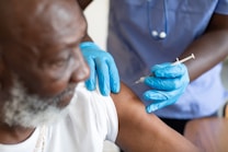 A healthcare worker wearing blue gloves is administering a vaccine to an elderly man. The scene focuses on the arm of the person receiving the injection and the hands of the healthcare worker handling the syringe.