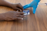 A person wearing blue medical gloves places a pulse oximeter on another person's finger. The device is used to measure oxygen saturation levels and is placed on a dark wooden table.
