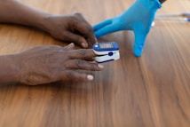 A person wearing blue medical gloves places a pulse oximeter on another person's finger. The device is used to measure oxygen saturation levels and is placed on a dark wooden table.