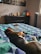 A dog is peacefully sleeping on a bed with a colorful geometric-patterned blanket. The bed is situated in a cozy bedroom with a dresser in the background. On top of the dresser, there are various decorative items, including a framed photograph, a white vase, a wooden hand sculpture, a metal tree sculpture, and a glowing orange lamp. A tower fan stands next to the window, and there are some dog toys on the bed.