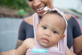 A smiling adult holds a baby close, both wearing soft pastel clothing. The baby looks curiously with big eyes and has a headband with a bow. The setting suggests a relaxed outdoor environment.