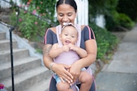 A woman is holding a smiling baby in her arms. The mother is wearing a black and pink outfit, and the baby has on a light purple outfit with a headband. They appear happy and relaxed, standing outside with greenery and stairs in the background.