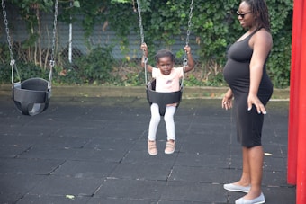 A young child sits in a swing on a playground, holding onto the chains. A pregnant woman stands nearby, smiling and looking at the child. The playground has rubber flooring and there is greenery in the background.
