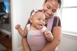 A woman lovingly holds a laughing baby in a soft fabric carrier. The baby wears a floral headband and appears to be enjoying the moment. Natural light pours in through a nearby window, adding a warm ambiance to the room.