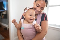 A woman lovingly holds a laughing baby in a soft fabric carrier. The baby wears a floral headband and appears to be enjoying the moment. Natural light pours in through a nearby window, adding a warm ambiance to the room.
