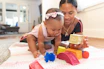 A smiling toddler playing with colorful blocks under the watchful eye of a warm, caring babysitter.