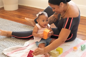 A woman and a baby are sitting on a colorful knitted blanket on a wooden floor. The baby is holding an orange block, while the woman is helping with other colorful blocks. Both appear engaged and smiling, suggesting a playful and nurturing interaction.