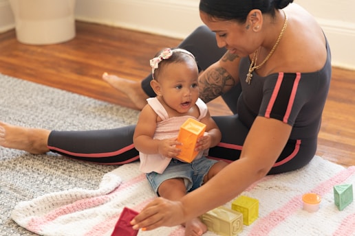 A woman and a baby are sitting on a colorful knitted blanket on a wooden floor. The baby is holding an orange block, while the woman is helping with other colorful blocks. Both appear engaged and smiling, suggesting a playful and nurturing interaction.