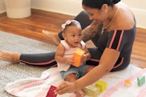 A woman and a baby are sitting on a colorful knitted blanket on a wooden floor. The baby is holding an orange block, while the woman is helping with other colorful blocks. Both appear engaged and smiling, suggesting a playful and nurturing interaction.