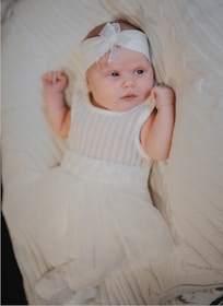 A baby wearing a white knitted outfit and matching headband lies comfortably on a soft, cream-colored blanket. The baby looks alert and curious, with a gentle and serene expression.