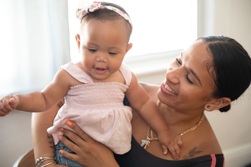 A joyful moment between a smiling woman and a baby, who is wearing a light pink dress with a matching headband. The woman, with tied-back hair, is holding the baby affectionately and looking at her with warmth. Sunlight filters through a nearby window, casting a soft glow over the scene.
