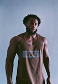 A man with a beard and afro hairstyle poses confidently wearing a brown tank top with the word 'UNRULY' printed on it. He stands against a plain background, giving a strong and composed presence.