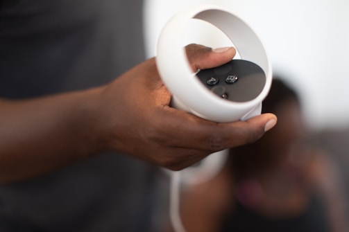 A person holds a VR controller with a white circular ring and black buttons. The focus is on the hand and controller, with a blurred background featuring another person.