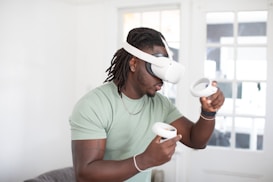 A person wearing a virtual reality headset, engaged with two handheld controllers. The background features a bright, softly lit room with a large window and a couch.