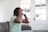 A person is sitting on a sofa wearing a virtual reality headset, their hands adjusting the device for a comfortable fit. The scene is set in a bright room with a large window featuring a door with multiple glass panes.