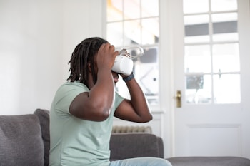 A person is sitting on a sofa wearing a virtual reality headset, their hands adjusting the device for a comfortable fit. The scene is set in a bright room with a large window featuring a door with multiple glass panes.