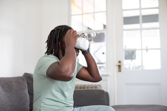 A person is sitting on a sofa wearing a virtual reality headset, their hands adjusting the device for a comfortable fit. The scene is set in a bright room with a large window featuring a door with multiple glass panes.