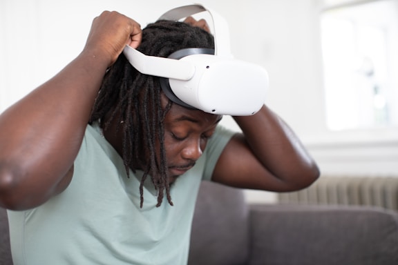 A person is putting on a virtual reality headset, adjusting it over their head while seated on a couch in a room with light-colored walls and a radiator in the background.