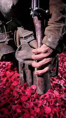 A person wearing worn military clothing, holding a vintage rifle, stands amidst a field of artificial red poppies. The poppies have black centers and create a striking contrast with the brown tones of the attire.