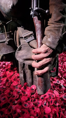 A person wearing worn military clothing, holding a vintage rifle, stands amidst a field of artificial red poppies. The poppies have black centers and create a striking contrast with the brown tones of the attire.