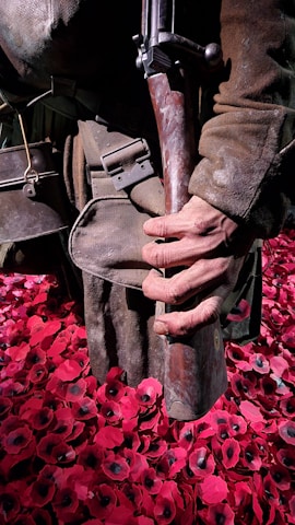 A person wearing worn military clothing, holding a vintage rifle, stands amidst a field of artificial red poppies. The poppies have black centers and create a striking contrast with the brown tones of the attire.