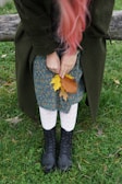 A person with long pink hair is holding autumn leaves while standing on grass. They are wearing a dark green coat, a floral-patterned dress, white textured tights, and black lace-up boots. The scene includes a wooden bench behind them.