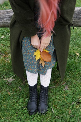 A person with long pink hair is holding autumn leaves while standing on grass. They are wearing a dark green coat, a floral-patterned dress, white textured tights, and black lace-up boots. The scene includes a wooden bench behind them.
