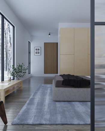 A modern bedroom with minimalistic design, featuring a platform bed with a dark blanket, a light-colored wooden floor, and large windows allowing natural light to illuminate the space. A small plant sits on a low wooden table next to the bed, and a soft gray rug extends across the floor. The walls are light, with a simple framed picture and a closed closet visible.