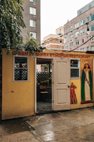A small, cozy Ethiopian restaurant with a welcoming open door. The exterior is decorated with a mural featuring traditional Ethiopian patterns and a figure in cultural attire. Surrounding buildings are visible in the background, suggesting an urban setting. There are string lights hanging above the entrance, adding a warm touch.