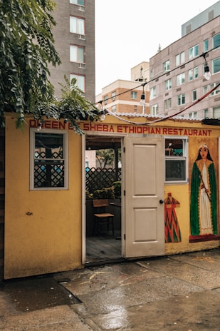 A small, cozy Ethiopian restaurant with a welcoming open door. The exterior is decorated with a mural featuring traditional Ethiopian patterns and a figure in cultural attire. Surrounding buildings are visible in the background, suggesting an urban setting. There are string lights hanging above the entrance, adding a warm touch.