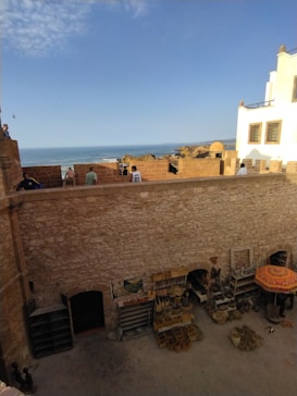 A stone courtyard with displays of traditional handmade baskets and woven items against a textured wall. Above the courtyard, people stand on a terrace, with a backdrop of an ocean meeting a clear blue sky. The terrace is bordered by stone walls, and a white building can be seen at the edge.