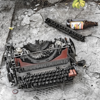 A broken typewriter is lying on cracked, weathered concrete. Beside it, there is an empty brown glass bottle labeled with a foreign language. A loose strip of metal and some scattered debris are also visible. A small yellow leaf is in the upper corner, adding a touch of color against the grayscale and muted tones.