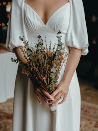 A person in a white dress holds a bouquet of dried flowers with eucalyptus and other foliage, focusing on their hands and the midsection of the dress. The lighting is soft, creating a gentle and serene atmosphere.
