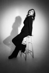A person in black clothing sits on a wooden stool in a minimalist setting. The image uses dramatic lighting to cast a shadow on the wall behind. The mood is contemplative, with the person looking upward, creating a contrast between light and dark areas.