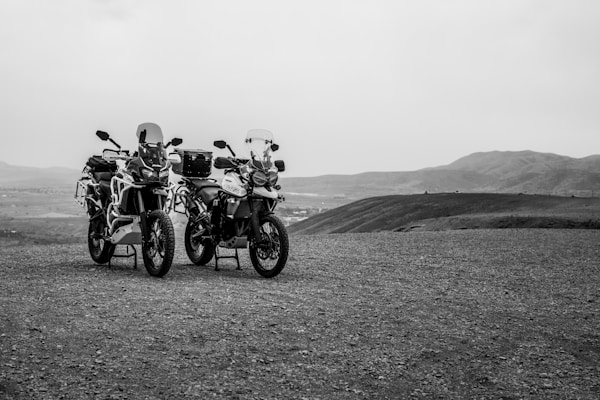 Two motorcycles are parked on a gravelly terrain with a vast landscape of hills and open sky in the background, creating a sense of adventure and exploration.