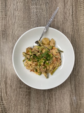 A white plate is filled with a dish that includes vegetables such as green beans and potatoes, and appears to be garnished with fresh herbs. The food is served with a spoon, and the setting features a wooden table surface.