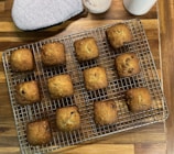 Twelve square, golden-brown muffins are arranged neatly on a cooling rack. They appear freshly baked with a slightly crispy exterior. The rack sits on a wooden surface alongside a grey oven mitt and some containers, possibly holding ingredients.
