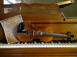 A violin rests on the keys of a wood-cased upright piano. Beside the violin, there is a sheet of music with a vintage appearance, showing handwritten musical notation. The background features a woven wooden cover of the piano's upper part.