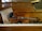 A violin rests on the keys of a wood-cased upright piano. Beside the violin, there is a sheet of music with a vintage appearance, showing handwritten musical notation. The background features a woven wooden cover of the piano's upper part.