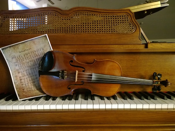 A violin rests on the keys of a wood-cased upright piano. Beside the violin, there is a sheet of music with a vintage appearance, showing handwritten musical notation. The background features a woven wooden cover of the piano's upper part.