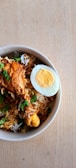 A bowl of biryani rice with pieces of chicken and a halved boiled egg. The dish is garnished with fresh green herbs and served in a white speckled bowl placed on a wooden surface.