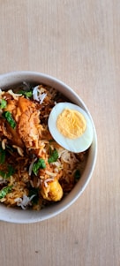 A bowl of biryani rice with pieces of chicken and a halved boiled egg. The dish is garnished with fresh green herbs and served in a white speckled bowl placed on a wooden surface.
