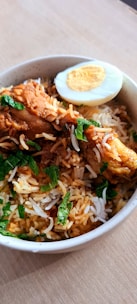 A bowl of biryani featuring spiced rice topped with pieces of chicken and garnished with fresh green herbs. A slice of hard-boiled egg is placed at the edge.