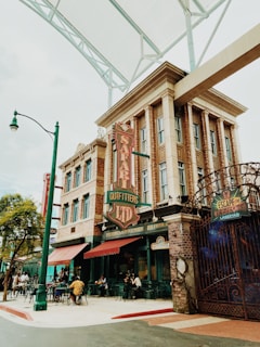 A bustling street scene featuring a vintage-style building with ornate architectural details and a retro 'Safari Outfitters Ltd' sign. People are seated at outdoor tables under green canopies, enjoying their time. The building has a classic design with brick and stone elements, and a large iron gate with a sign for 'Escape the RE'. Trees and streetlights line the sidewalk.