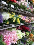 A variety of colorful artificial flowers are displayed on metal racks. The flowers include lilacs, roses, lilies, and tulips in shades of pink, yellow, red, purple, and white. Price tags are visible above the flowers, indicating a retail setting.