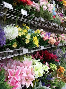 A variety of colorful artificial flowers are displayed on metal racks. The flowers include lilacs, roses, lilies, and tulips in shades of pink, yellow, red, purple, and white. Price tags are visible above the flowers, indicating a retail setting.
