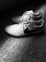 A pair of white soccer cleats with black accents resting on a textured carpet, illuminated by light casting shadows.