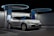 A sleek, white electric vehicle is parked at a futuristic-looking charging station. The environment has a modern design with circular overhead structures emitting blue light, enhancing the technological ambiance. The car's headlights are distinct, and there's a digital display screen nearby indicating a status related to charging.