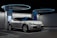 A sleek, white electric vehicle is parked at a futuristic-looking charging station. The environment has a modern design with circular overhead structures emitting blue light, enhancing the technological ambiance. The car's headlights are distinct, and there's a digital display screen nearby indicating a status related to charging.