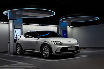 A sleek, white electric vehicle is parked at a futuristic-looking charging station. The environment has a modern design with circular overhead structures emitting blue light, enhancing the technological ambiance. The car's headlights are distinct, and there's a digital display screen nearby indicating a status related to charging.